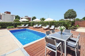 A swimming pool with lounge chairs and umbrellas, a wooden deck, and a table with chairs. - Villa Periwinkle (Paphos)