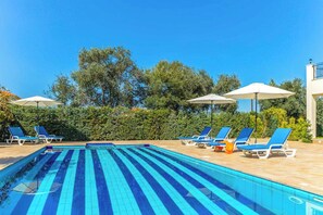 Une piscine avec des lignes de démarcation bleues, entourée de chaises longues et de parasols, avec un ciel dégagé et des arbres en arrière-plan.