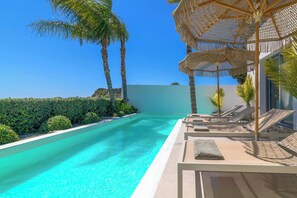 A clear blue pool with lounge chairs and umbrellas, surrounded by palm trees and greenery.