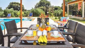 A dining table set with breakfast items, surrounded by wicker chairs, with a pool and greenery in the background. - Villa Carreta (Corfu)