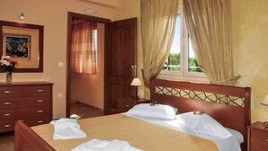 A hotel room with a bed, a wooden dresser, a nightstand, a lamp, and a window with curtains. - Villa Carreta (Corfu)