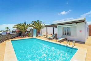 Piscina con sillas de descanso y una zona cubierta, rodeada de palmeras y un muro de piedra.