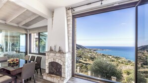 A dining area with a stone fireplace and a large window overlooking a coastal landscape. - Pomos View (Pomos)