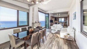 A spacious living area with a dining table, comfortable seating, a fireplace, and a large window offering a view of the ocean. - Pomos View (Pomos)