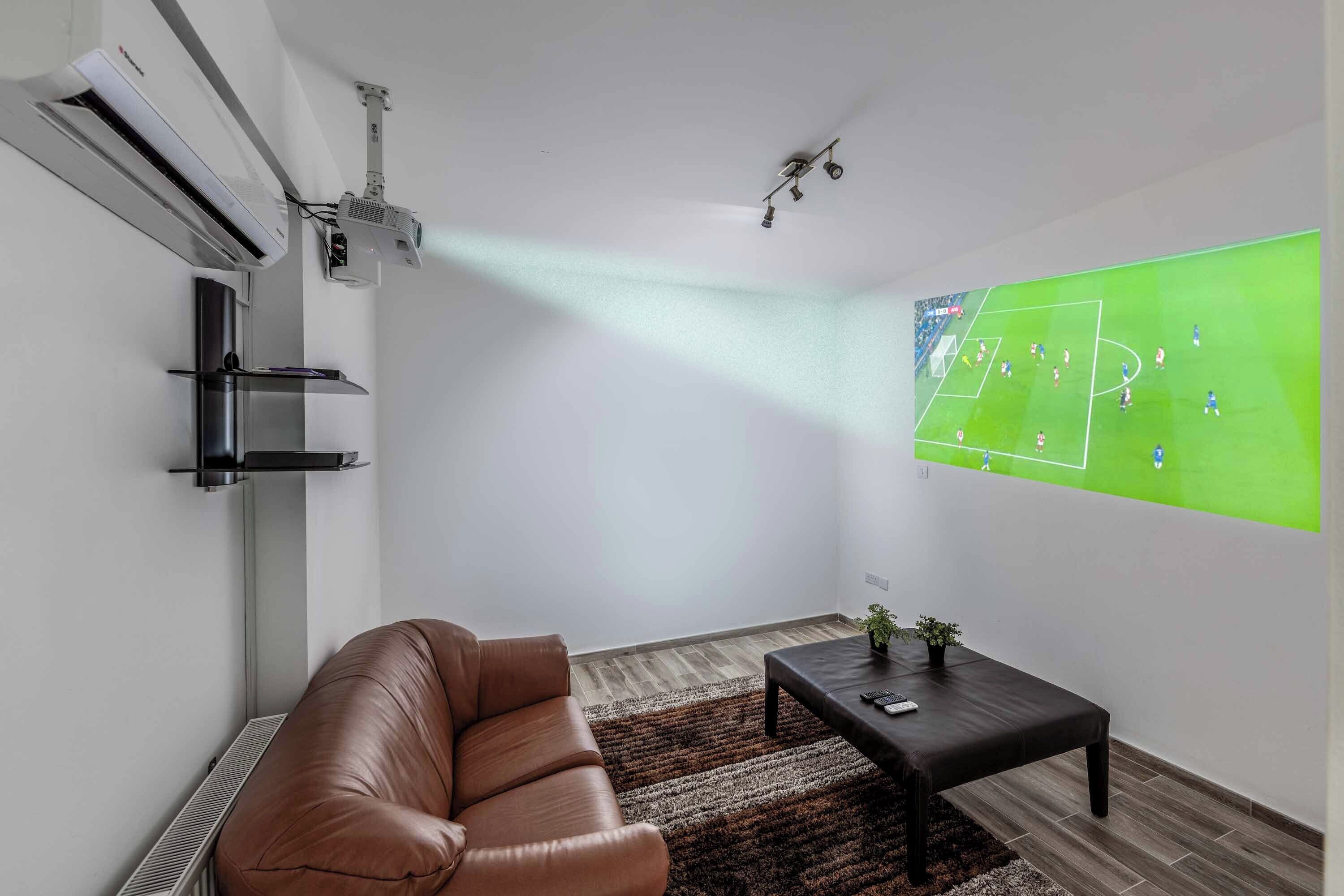 A modern living room with a large wall-mounted TV displaying a soccer game, a leather sofa, a coffee table, and a small plant.