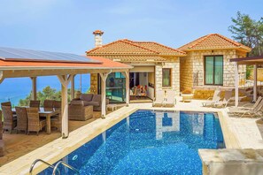 A poolside area with a clear view of the sea, a covered seating area, and a stone building with a tiled roof.