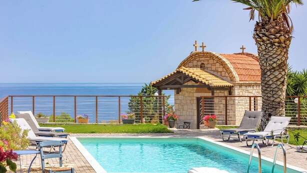 Zona de piscina con sillas de descanso, un pequeño edificio con una cruz en la parte superior y vistas al mar.
