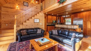 A log cabin interior with a wooden staircase, two leather sofas, a coffee table, and a kitchen area. - Morris Cabin (Boone)