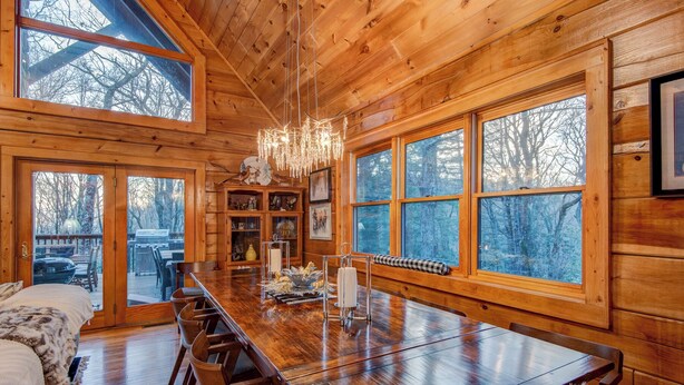 A wooden cabin interior with a large dining table, multiple chairs, a chandelier, and large windows offering views of the surrounding forest. - Blueberry Hills (Boone)