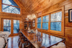 A wooden cabin interior with a large dining table, multiple chairs, a chandelier, and large windows offering views of the surrounding forest. - Blueberry Hills (Boone)