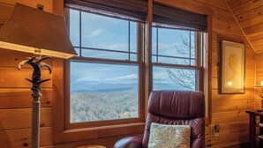 A wooden cabin room with a large window, a leather chair with a map cushion, a lamp, and a framed picture on the wall. - Deer Moments (Banner Elk)