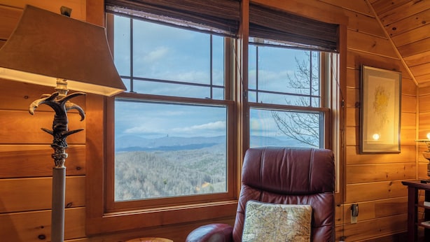 A wooden cabin room with a large window, a leather chair with a map cushion, a lamp, and a framed picture on the wall. - Deer Moments (Banner Elk)