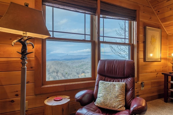 A wooden cabin room with a large window, a leather chair with a map cushion, a lamp, and a framed picture on the wall. - Deer Moments (Banner Elk)