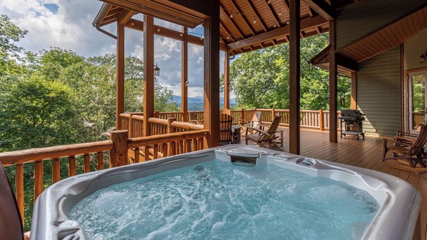 Una terraza de madera con un jacuzzi, muebles de exterior y vistas a árboles y montañas.