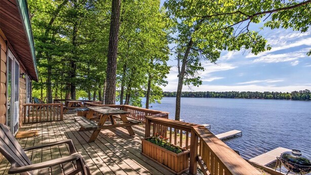 A wooden deck with outdoor furniture, a grill, and a view of a lake surrounded by trees.
