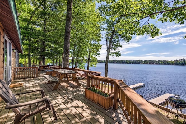 Una terraza de madera con mobiliario exterior, una parrilla y vistas a un lago rodeado de árboles.