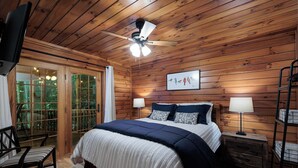 Ein gemütliches Holzhaus-Zimmer mit einem Bett, zwei Nachttischlampen, einem Deckenventilator und einem Fenster mit Blick auf den Wald.