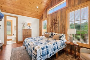 A bedroom with a large bed, wooden walls, a window with blinds, a bedside table with a lamp, and a view of greenery outside. - Banner Sky (Banner Elk)