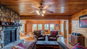 A cozy cabin interior with a stone fireplace, wooden walls, a ceiling fan, and a large window offering a view of the outdoors. - Chestnut Lodge (Boone)