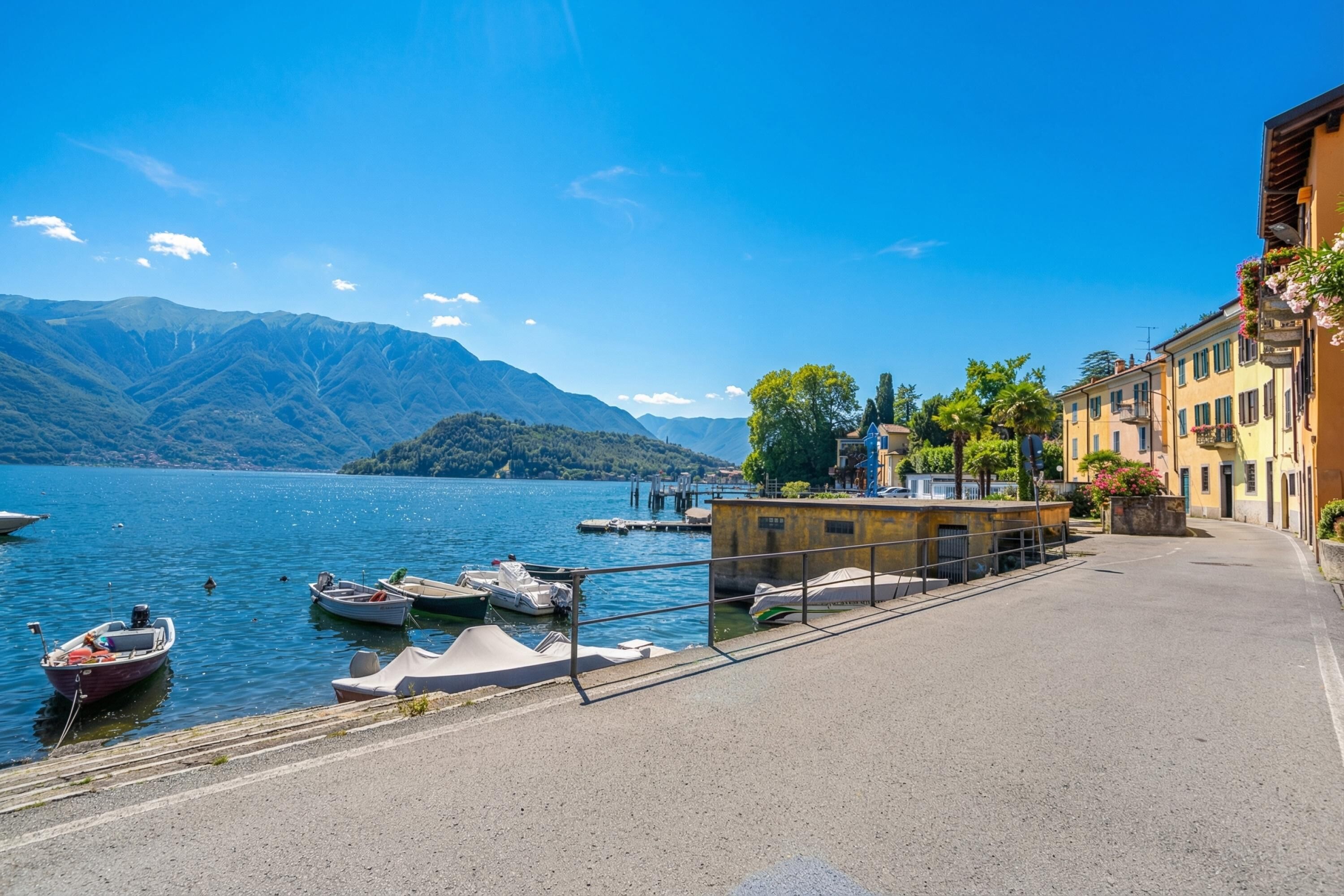 A lakeside street with boats docked, a building with a balcony, and mountains in the background.