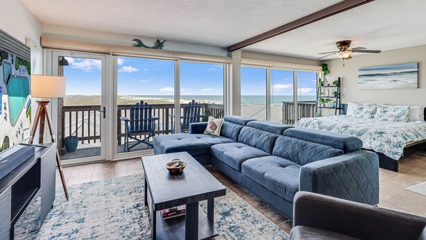 A spacious living area with a large sofa, a coffee table, and a television. There is a balcony with a view of the ocean. - Gulfstream Condos Unit 603 | Beautiful Gulf Views | Pool, Hot Tub, BBQ (Corpus Christi)