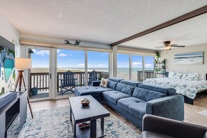 A spacious living area with a large sofa, a coffee table, and a television. There is a balcony with a view of the ocean.