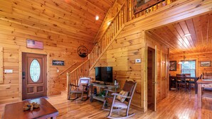 Interior de una cabaña de madera con comedor, televisor, sillones mecedores y una escalera.