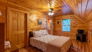 Un dormitorio de cabaña de madera con una cama, un ventilador de techo, una ventana y una mesita.