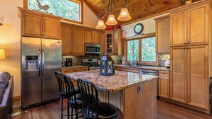 A kitchen with wooden cabinets, a stainless steel refrigerator, a granite countertop, and a central island with bar stools. - Another Point of View (Blowing Rock)