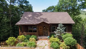 A wooden cabin with a stone entrance, surrounded by lush greenery and a stone pathway leading to the door. - Another Point of View (Blowing Rock)