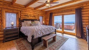 Une chambre située dans un chalet en rondins, avec un grand lit, une tête de lit en bois, une table de chevet, un banc et une vue sur l’extérieur.