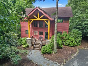 Une maison à deux étages, à l’extérieur rouge et aux accents jaunes, entourée d’une végétation luxuriante.
