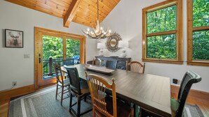 A dining room with a wooden table, chairs, a chandelier, and large windows with a view of greenery. - Muir Lodge (Blowing Rock)