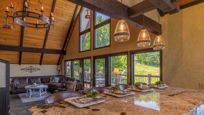 A spacious living area with a large granite island, multiple seating options, and large windows offering views of the outdoors. - Halftime Haus (Banner Elk)