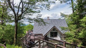 A cabin with a stone chimney, surrounded by lush greenery and a wooden deck. - Halftime Haus (Banner Elk)