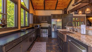A spacious kitchen with dark wood cabinets, stainless steel appliances, and large windows offering a view of greenery. - Halftime Haus (Banner Elk)