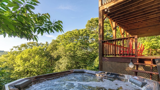 A hot tub with a wooden deck and red chairs, surrounded by lush greenery. - Whispering Ridge Retreat (Boone)