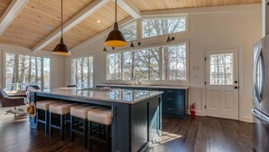 A spacious kitchen with a large island, bar stools, pendant lighting, and large windows offering views of trees.
