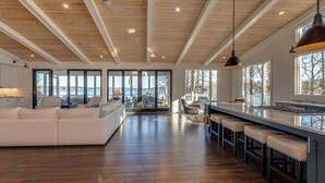 A spacious living area with a large white sofa, a kitchen bar with stools, and a view of the outdoors through large windows.