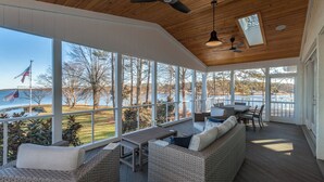 A covered outdoor deck with wicker furniture, large windows, and a view of a lake.