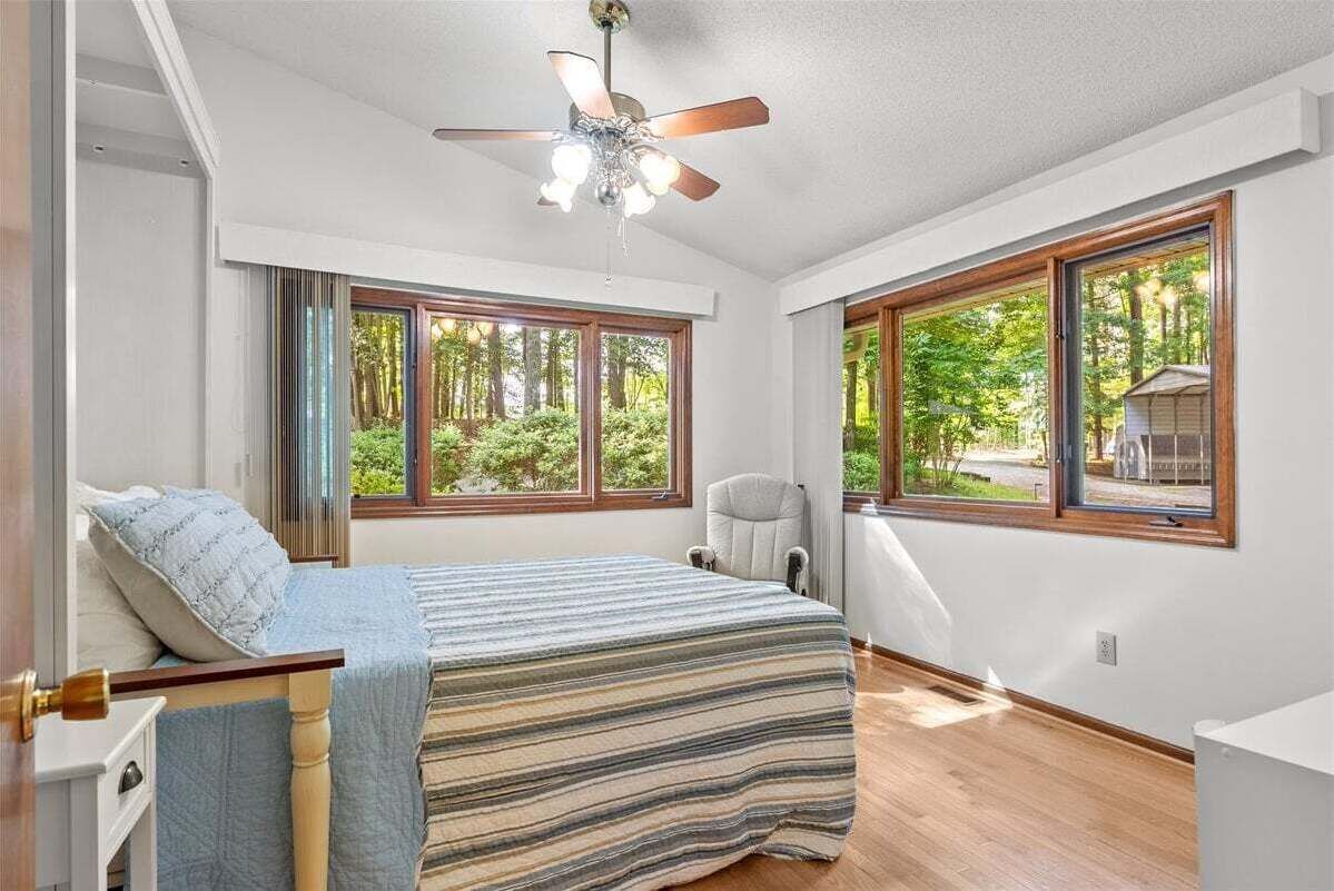 A bedroom with a bed, a chair, a ceiling fan, and large windows with a view of trees.
