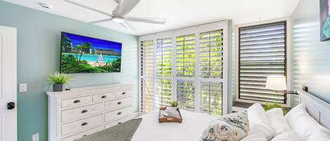 Un dormitorio con cama, ventilador de techo, televisión, ventana con persianas y mesita de noche con una planta.