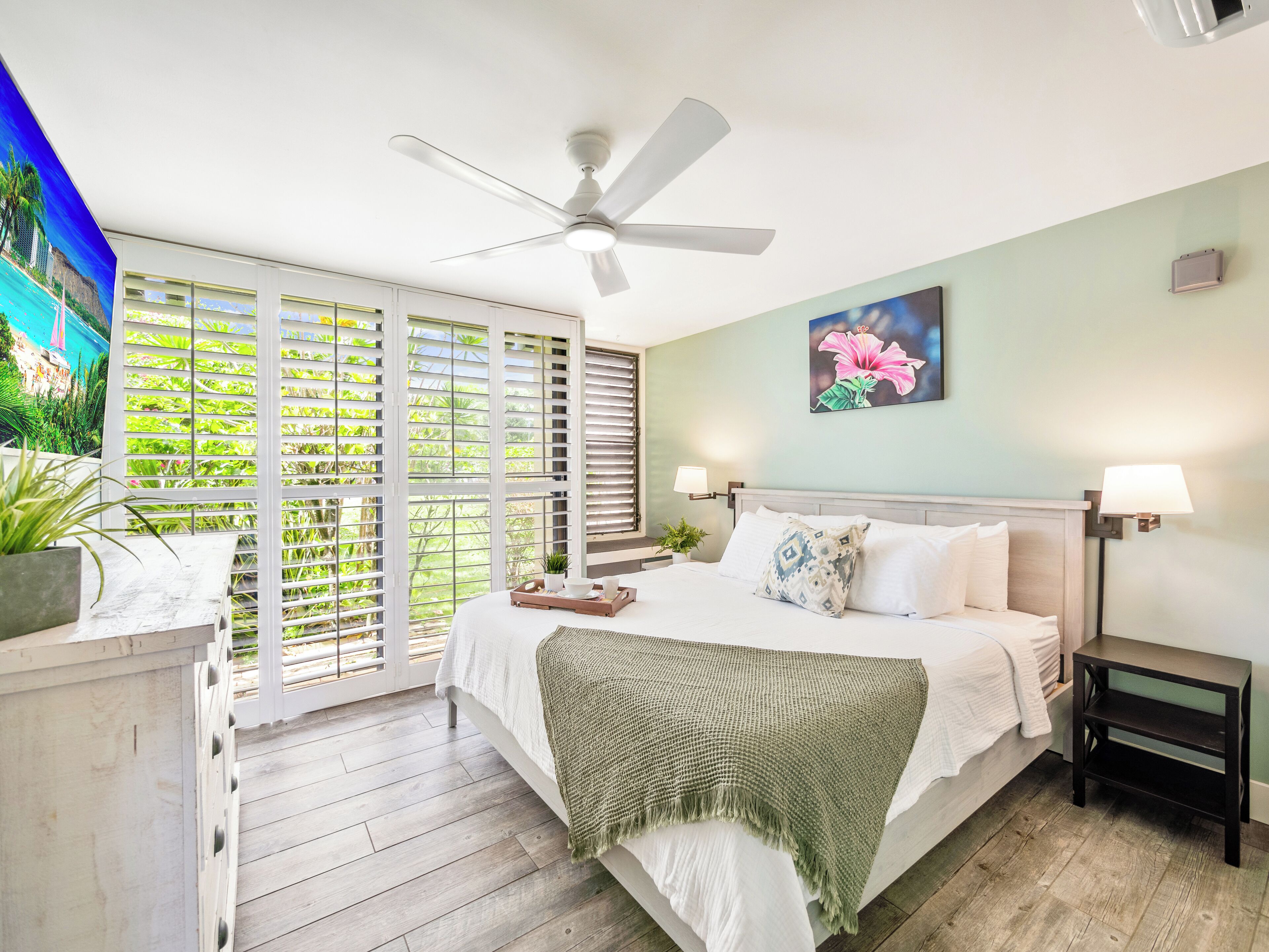 A bedroom with a bed, a ceiling fan, a window with blinds, a nightstand, and a wall-mounted lamp.