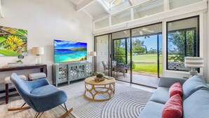 A spacious living room with a large flat-screen TV, comfortable seating, and a view of a lush garden through large sliding glass doors.