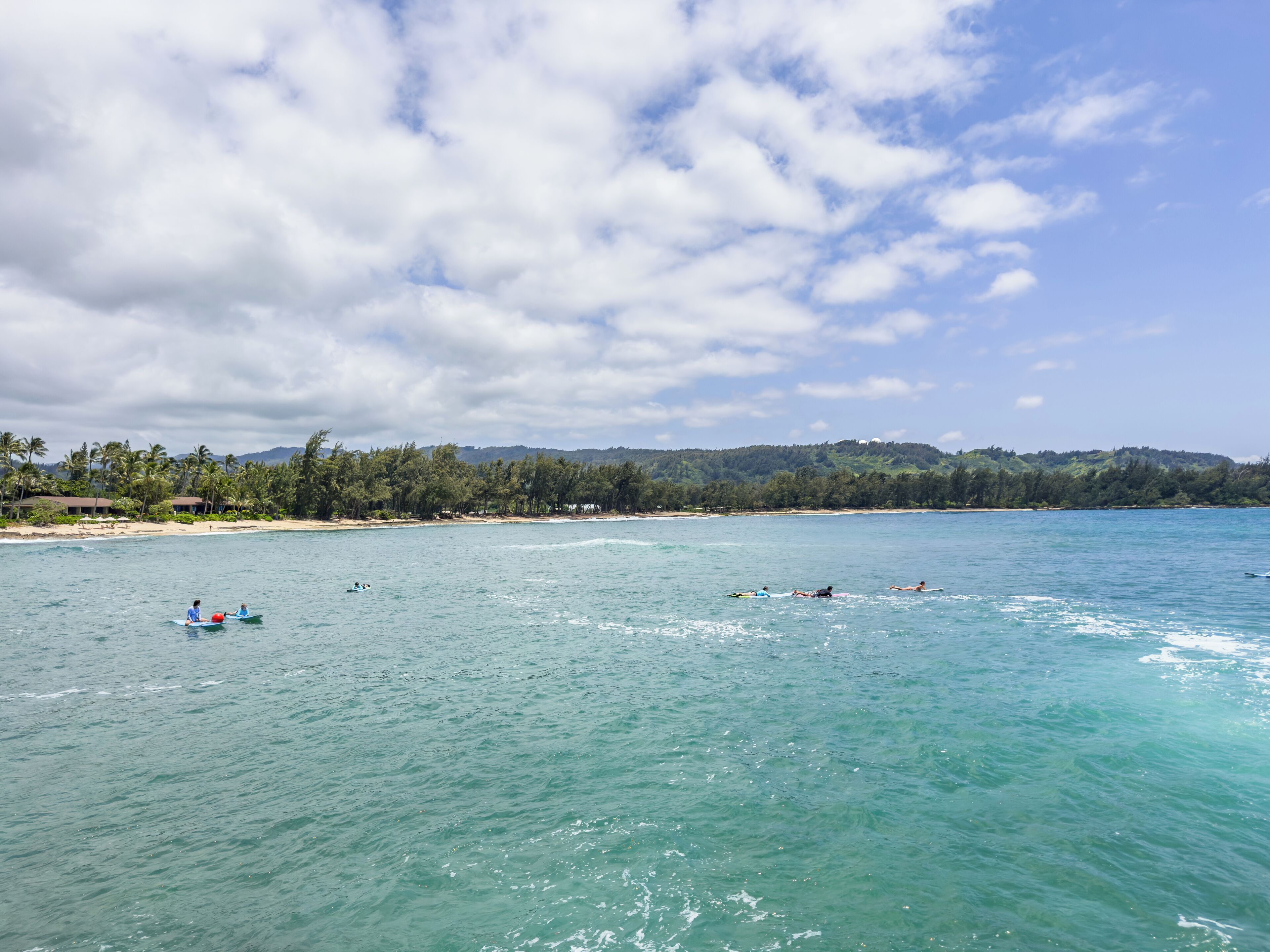 Une plage bordée d’une eau turquoise et limpide, de palmiers et d’un paysage vallonné en arrière-plan.