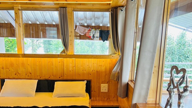 A wooden cabin room with a bed, a bench, and a view of the outdoors through large windows. - Snooze Cottage (Dhari)