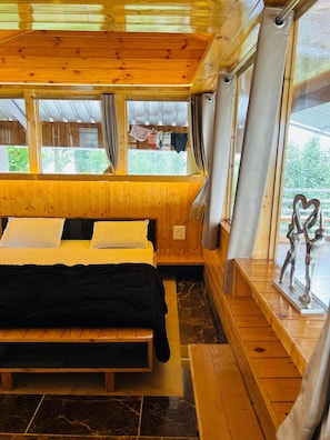 A wooden cabin room with a bed, a bench, and a view of the outdoors through large windows. - Snooze Cottage (Dhari)