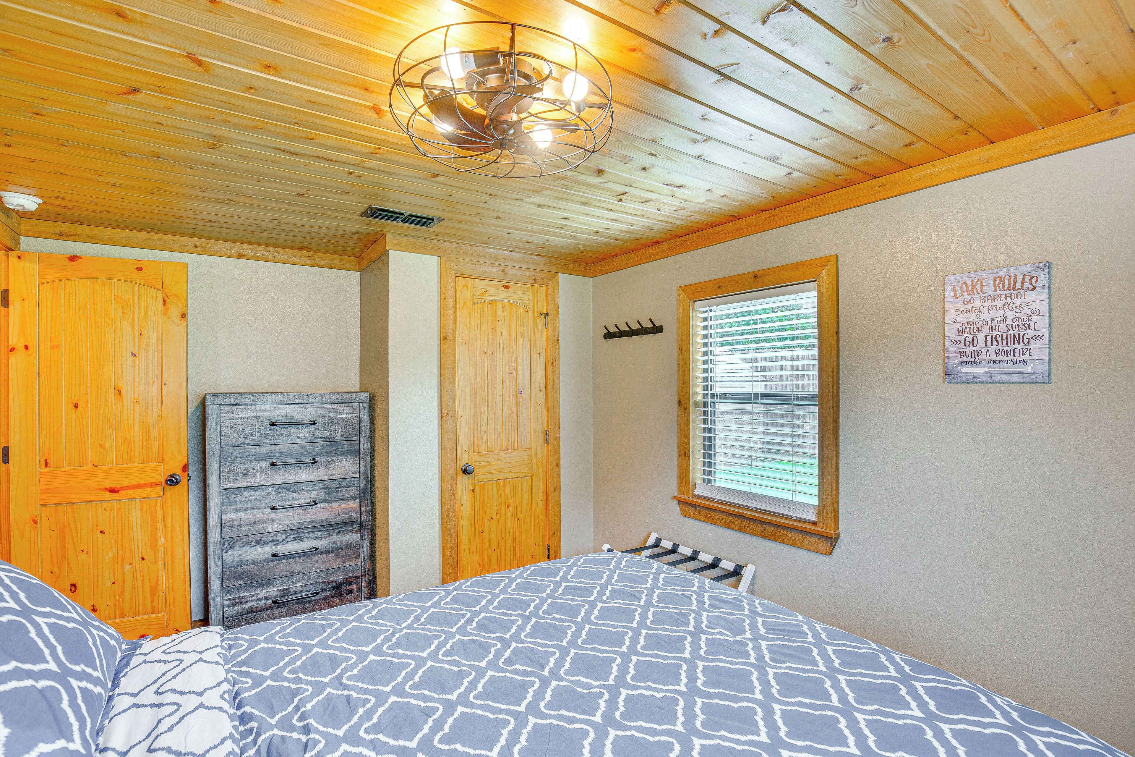 A bedroom with a wooden ceiling, a bed with a patterned bedspread, a dresser, and a window with blinds.