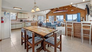 Een ruime woonkamer met een houten plafond, een eettafel met stoelen, een keuken en een groot raam met gordijnen.