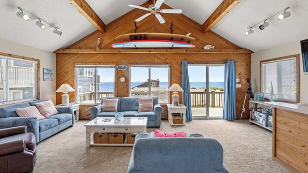 Een ruime woonkamer met een hoge plafond, houten balken, een plafondventilator en een decoratie van een surfplank.